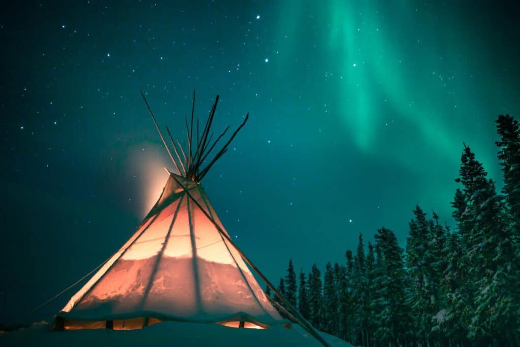 Worms eye view shot of a Tipi under a wintery landscape and a starry night sky with an aurora.