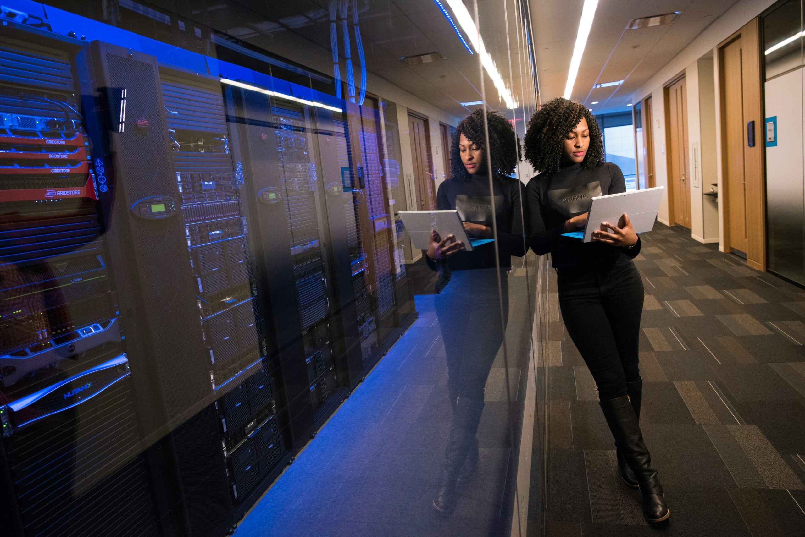 A person holding a laptop beside a room of server racks.